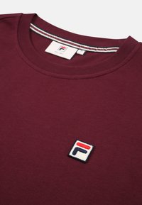 Camiseta burdeos de mezcla de algodón con cuello redondo y mangas cortas. Presenta un parche de logo bordado en rojo, blanco y azul en el pecho.