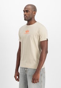 Camiseta de algodón beige con mangas cortas, cuello redondo y logo gráfico rojo en el pecho; combinada con jeans de mezclilla gris claro.