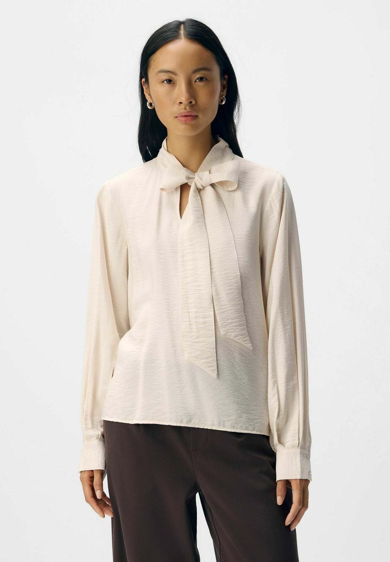 Cremfarbene Bluse mit einer Schleifen-Detail am Kragen, langen Ärmeln und einem strukturierten Stoff. Kombiniert mit dunklen Hosen.
