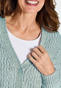 Helleblaue Strickjacke mit strukturiertem Muster, mit Knopfleiste und V-Ausschnitt, über einem weißen Hemd getragen.