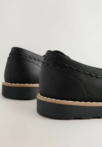 Chaussures en cuir noir à enfiler avec un design festonné, dos arrondi et une semelle beige avec des coutures blanches.