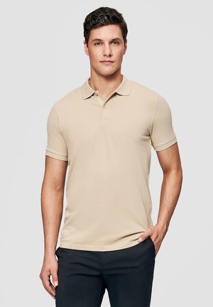 Mężczyzna o krótkich ciemnych włosach, ubrany w beżową koszulkę polo z krótkim rękawem i ciemne spodnie, stojący z jedną ręką w kieszeni na szarym tle.