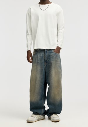 Personne portant une chemise blanche à manches longues, un jean bleu ample et délavé, des baskets blanches et un collier fin en chaîne, debout devant un fond uni.