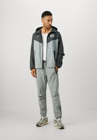 Grå og svart to-farget vindjakke med hette, glidelåslommer og logo. Sammen med lysegrå bukser og mønstrede sneakers.