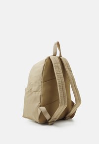 Mochila de nylon beige, con correas de malla acolchadas, forma redondeada y bolsillos laterales. Textura suave con un acabado mate y sutilezas en los detalles de costura.