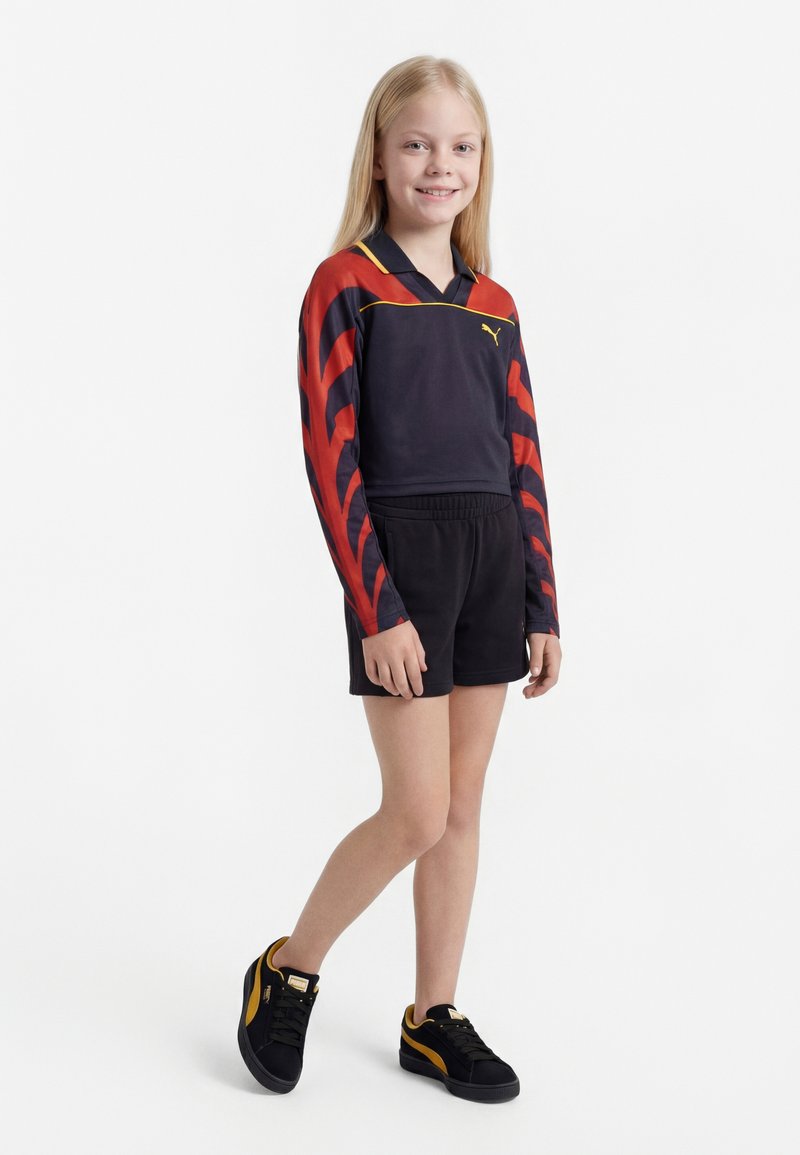 Deklica stoji in se smeji, oblečena v črno-rdečo majico z dolgimi rokavi, črne kratke hlače in črne superge z rumenimi poudarki.