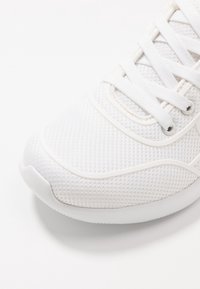 Gros plan sur l'empeigne d'une basket blanche en tissu en maille, avec des lacets et une semelle en caoutchouc sur fond blanc.