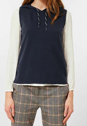 Femme portant un sweat à capuche sans manches bleu marine sur un chemisier blanc à manches longues et un pantalon à carreaux avec des lignes roses et jaunes.