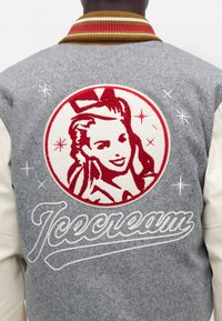 Vue de dos d'une personne portant une veste grise et blanche avec un visage de femme rétro brodé en rouge et blanc et le mot « Icecream » dans le dos.