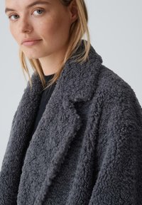 Gray fleece coat with a textured, shaggy exterior. It features a shawl collar and is worn over a black top, showcasing a soft, cozy material.