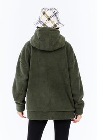 Hoodie en polaire verte surdimensionnée avec une finition texturée, dotée d'une grande capuche et de manches tombantes, associée à un chapeau seau à motifs.