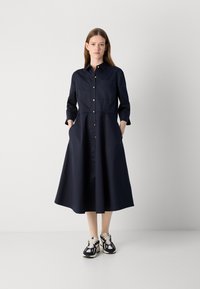 Marineblauwe hemdjurk met een knopenlijst aan de voorkant, driekwart mouwen, een borstzak en zijzakken; gecombineerd met donkere sneakers. Glad materiaal, A-lijn vorm.