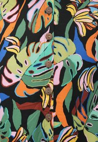 Tissu multicolore avec un motif de feuilles tropicales en vert, orange et rose. Présente des boutons marron alignés au centre.