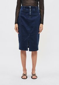 Tamsiai mėlynos spalvos džinsinis pieštuko formos sijonas su užtrauktuko uždarymu, priekiu su skeltuku ir juosmens detalėmis. Modelis vilki juodus butus be kulnų.