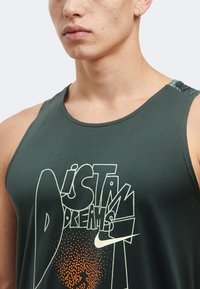 Νέος άνδρας που φοράει σκούχο αμάνικο μπλουζάκι Nike με γραφικά «Just Do Your Dreams» και πορτοκαλί πουά μοτίβο, στέκεται μπροστά σε απλό φόντο.