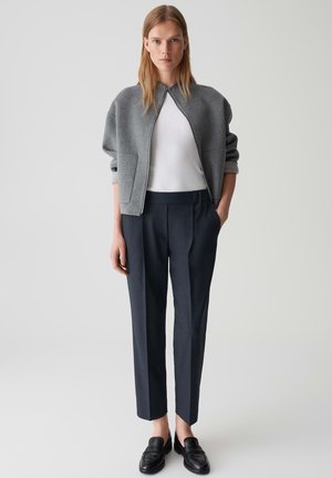 Grau geschnittene Jacke mit Reißverschluss, getragen über einem weißen Oberteil, kombiniert mit navyfarbigen Hosen und schwarzen Slippern. Glatte Texturen und taillierte Passform.