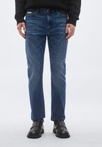 Μπλε τζιν παντελόνι με ίσια γραμμή, διακριτική �ξεθώριαση, πέντε τσέπες και κλείσιμο με μεταλλικό κουμπί, συνδυασμένο με μαύρα παπούτσια.