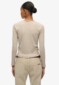 Haut beige à manches longues avec texture côtelée, coupe ajustée et col rond. Porté avec un pantalon clair, taille haute, doté de deux poches arrière.
