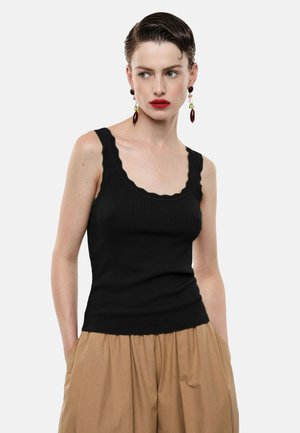 Donna con capelli tirati indietro che indossa una canotta nera con bordi smerlati, pantaloni beige a vita alta, rossetto rosso acceso e orecchini pendenti.