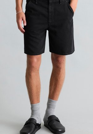 Mężczyzna w czarnych spodenkach do kolan, szarych skarpetach do kostek, czarnych butach wsuwanych, stoi z rękami w kieszeniach na jednolitym tle.