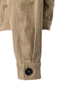 Tarnfarbene Cordjacke mit gerippter Textur. Verfügt über einen schwarzen Knopf am Ärmelbündchen, der detaillierte Nähte und eine strukturierte Passform zeigt.