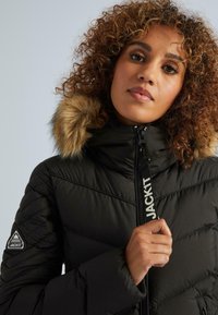 Sieviete ar cirtainiem matiem, ģērbusies melnā pufīgā jakā ar kažokādas apmali ap kapuci, turot rokas rāvējslēdzēja vilkšanas elementu ar uzrakstu "JACK1T".