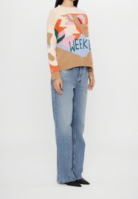 Sweter dzianinowy z wielokolorowymi abstrakcyjnymi wzorami, z napisem "WEEKEND". Połączony z jasnoniebieskimi jeansami i czarnymi butami z zaostrzonym noskiem.
