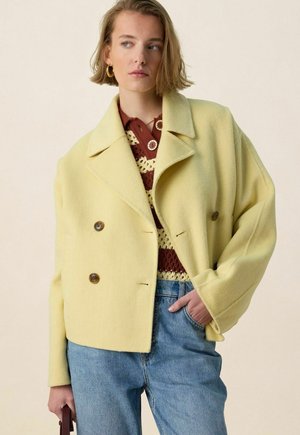 Femme portant une veste en laine jaune pâle à double boutonnage sur un top en crochet marron et crème, un jean bleu et des boucles d'oreilles créoles dorées.