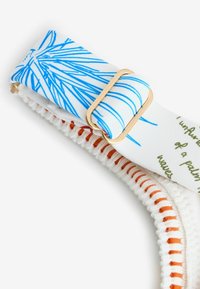 Modrý a biely vzorovaný nastaviteľný remienok so zlatou prackou pripevnený k bielej látke s oranžovým prešívaním a čiastočným zeleným textom.