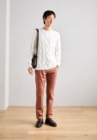 Valkoinen verkkoneule, vaaleanruskeat chinot ja mustat loaferit. Mallilla on pieni musta olkalaukku. Neutraali tausta puulattialla.