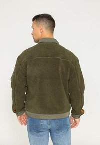 Zielona oliwkowa kurtka z sherpy, charakteryzująca się miękką, teksturowaną powierzchnią, prążkowanymi mankietami z pomarańczowymi akcentami oraz usztywnionym kołnierzem.