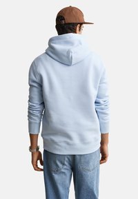 Homme portant un sweat à capuche bleu clair, jean bleu, casquette marron et montre-bracelet, vu de dos sur un fond uni.