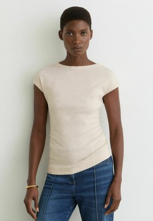 Vrouw met kort haar die een beige T-shirt met korte mouwen en blauwe spijkerbroek draagt, staand tegen een witte muur, kijkt recht naar voren.