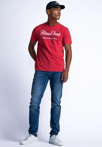 Red cotton t-shirt with "Petrol Ind." text, short sleeves, paired with blue denim jeans and white sneakers. Simple cap completes the look.