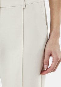 Lichtbeige op maat gemaakte broek met een gladde textuur, voorzien van een subtiele vouw aan de voorkant en een zijzak. Close-up van een hand op de stof.