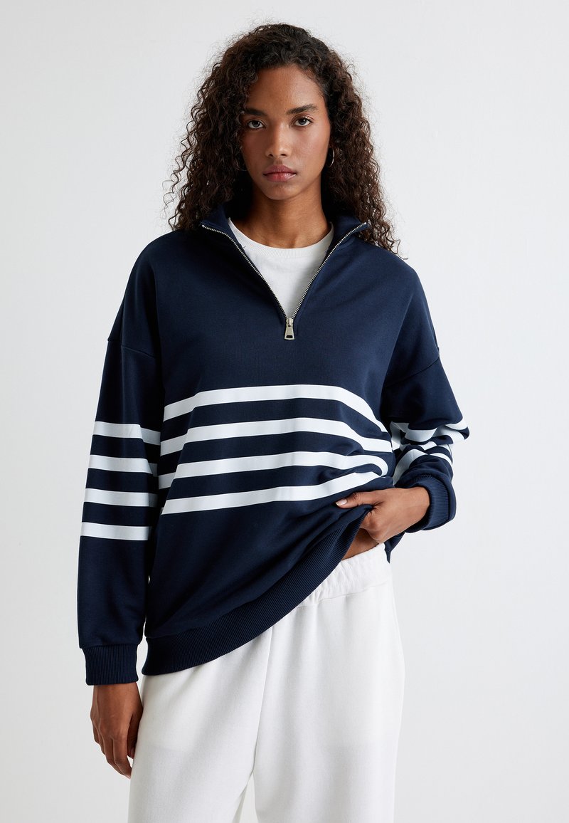 Sudadera con cremallera en color marino con rayas horizontales blancas; corte holgado, puños acanalados y textura suave; lleva puesta encima de una camiseta blanca con pantalones relajados.