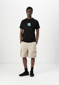 Sort t-shirt med blå blomsterbroderi på brystet, parret med beige cargo shorts med sidelommer og sorte sneakers.