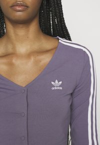 Lila långärmad topp med v-ringning, med vita sidostripes och en liten vit Adidas-logotyp. Knappdetalj i mitten fram.