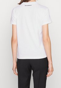 Camiseta de algodón blanca con cuello redondo y mangas cortas. Logo "KARL LAGERFELD" impreso en negro en la parte superior de la espalda. Diseño minimalista.