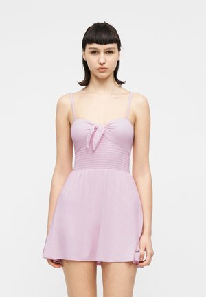 Fiatal nő rövid fekete hajjal, világos lila ujjatlan ruhát visel, amelynek elülső részén kötődés található, fehér háttér előtt áll.