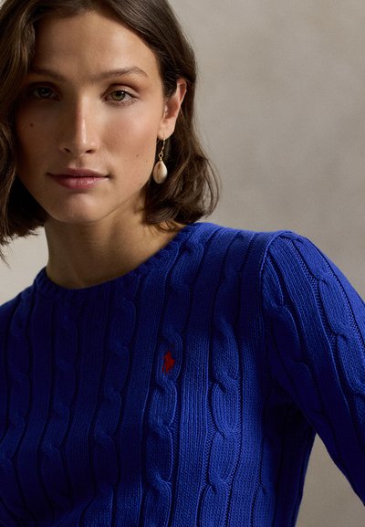 Kongelig blå strikkegenser med kabelmønster og liten rød logo. Modellen har en droppende øredobb, med myk, naturlig sminke og kort hår.