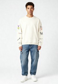 Krém színű pulóver színes hímzett részletekkel az ujjain, párosítva kék cargo farmerral és fehér tornacipővel. Lazább fazon és dizájn.