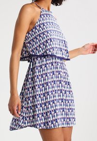 Kvinna iklädd en ärmlös klänning med geometriska mönster i blått, vitt och lila, med lager på toppen och utsvängd kjol, framför en neutral bakgrund.