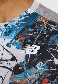 Nærbillede af en farverig sportstrøje med abstrakt blå, sort, hvid og orange malingprikke samt en hvid New Balance-logo på brystet.