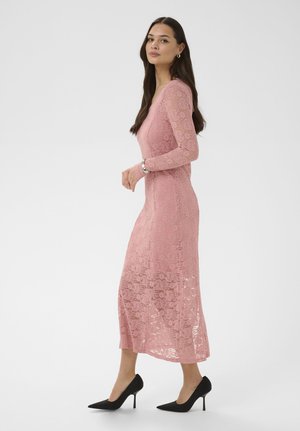 Femme aux cheveux longs et bruns, portant une robe midi en dentelle rose à manches longues et des escarpins noirs, debout de profil sur un fond blanc.