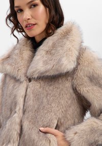 Abrigo de piel sintética en beige suave con un amplio cuello, que presenta una textura aterciopelada y un frente de doble botonadura, elegantemente cálido para climas más frescos.