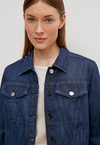 Veste en denim bleu foncé avec des détails en acier inoxydable, dotée de fermetures à boutons, de deux poches poitrine et d'un design de col classique.