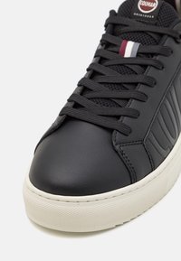 Svart lädersneaker med strukturerade paneler, platta svarta snören, vit sula och en liten accent i rött, vitt och blått nära tungan.