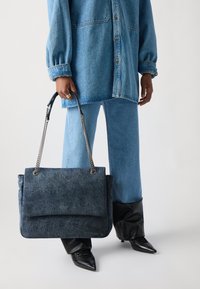 Bolso tote de denim en azul oscuro con cierre de solapa y asa de cadena. Textura suave con costuras visibles y detalle del logo en la parte delantera.