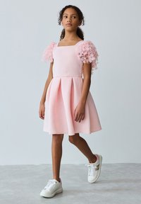 Tyttö kävelee vaaleanharmaata, yksiväristä taustaa vasten pukeutuneena vaaleanpunaiseen mekkoon, jossa on kukalliset, tylliset hihat, ja valkoiset lenkkarit.